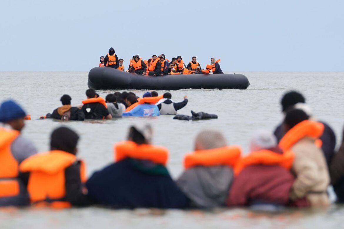 Crowded inflatable dinghy with many people wearing bright orange life jackets on the sea.