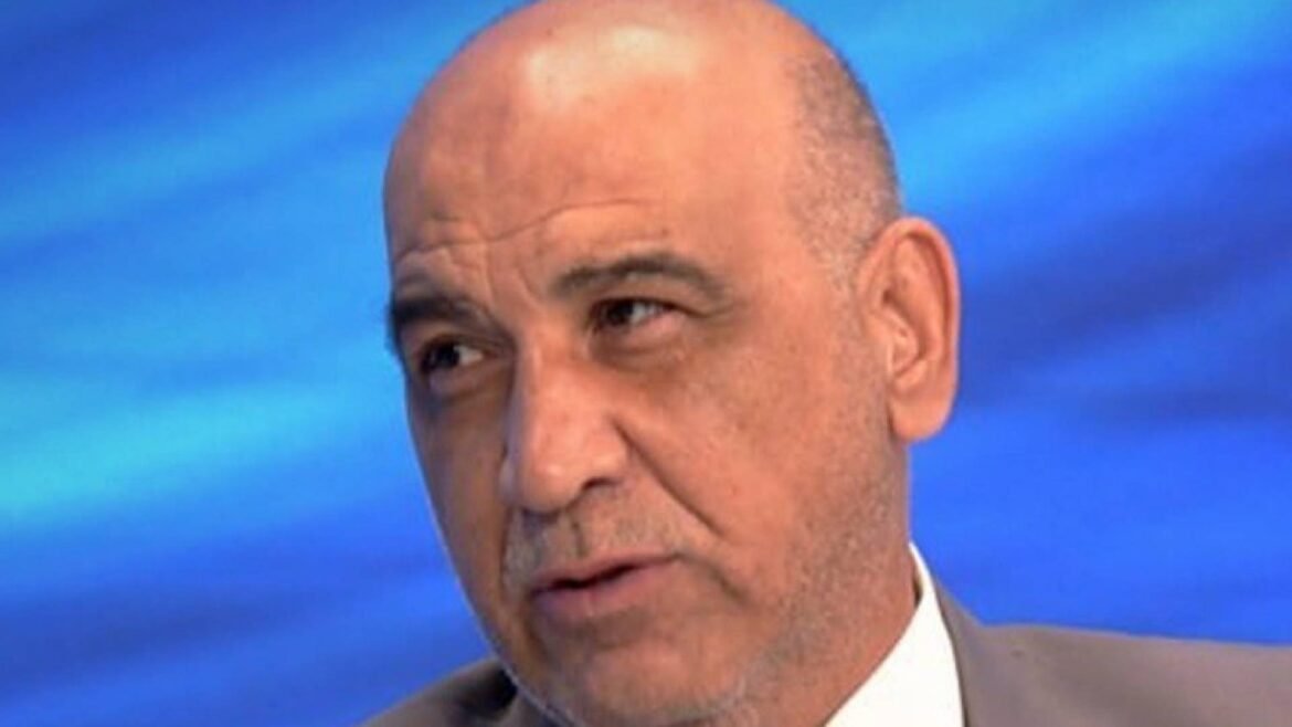 Close-up of a middle-aged man with a shaved head and light stubble, wearing a suit, speaking on a blue TV studio backdrop