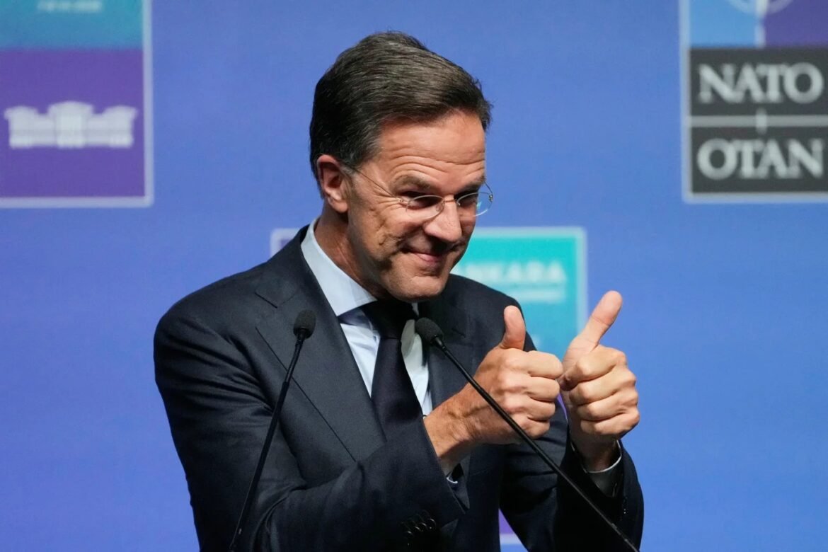 Man in a dark suit at a podium giving two thumbs-up during a press conference with blue backdrop and microphones in front of him.