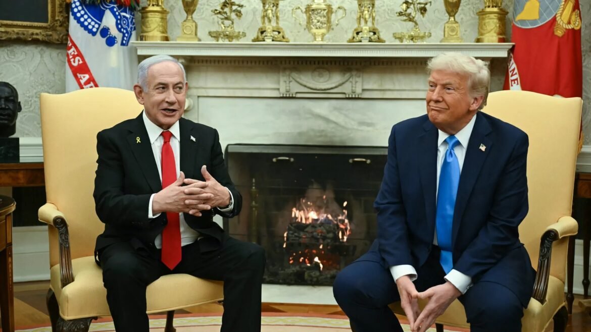 Two suited men sit in yellow armchairs beside a fireplace, engaging in conversation, with gold trophies on the mantel and flags in the background.
