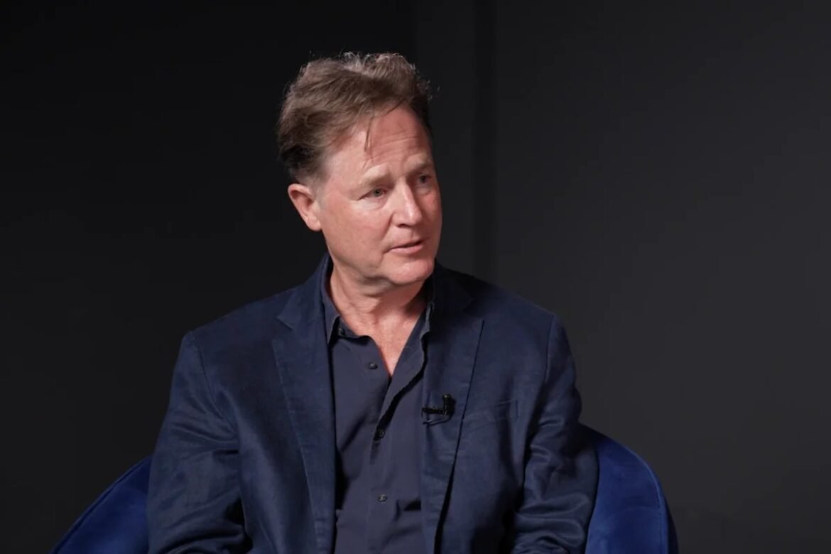 Man in a navy suit sits on a blue chair, speaking into a wireless mic during an interview or panel discussion in a dark studio.