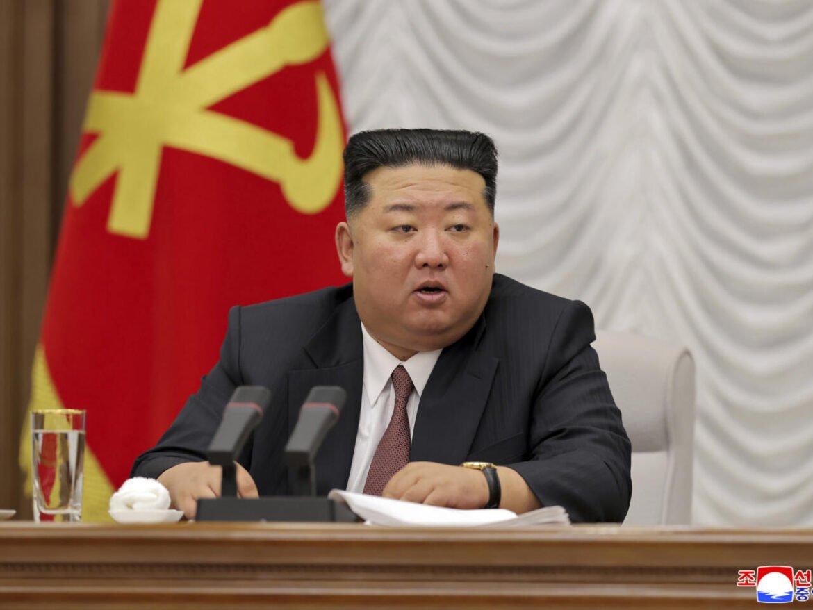 North Korean leader Kim Jong-un seated at a desk with microphones, speaking in a formal setting with a red flag in the background.