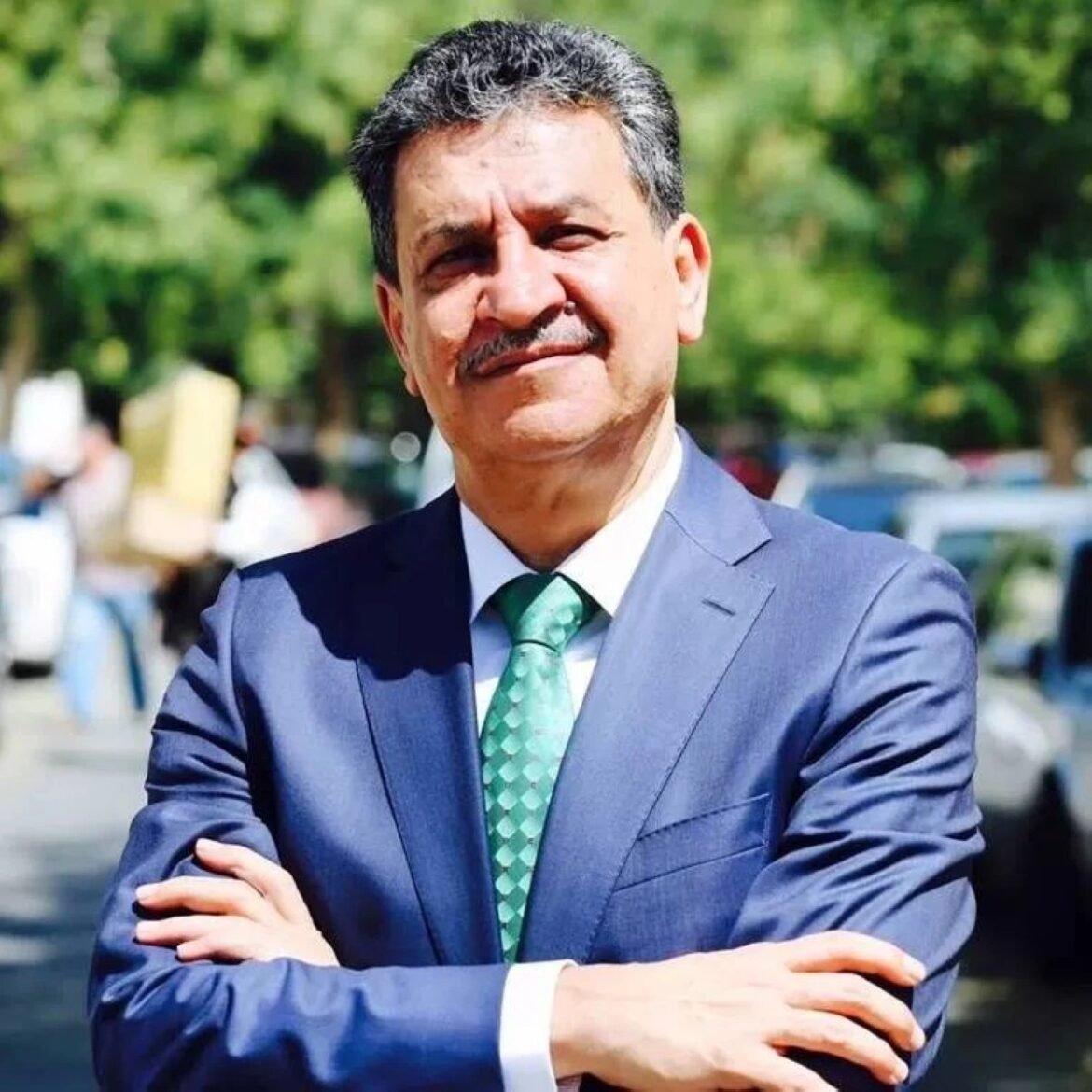 Smiling man in a blue suit and green tie posing with arms crossed outdoors.
