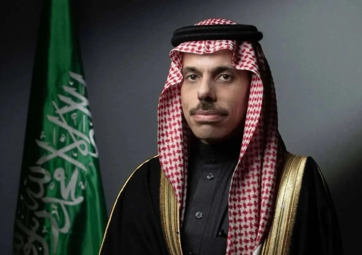 Portrait of a man in a red-and-white keffiyeh and black thobe with a gold-trimmed bisht, beside the Saudi flag.