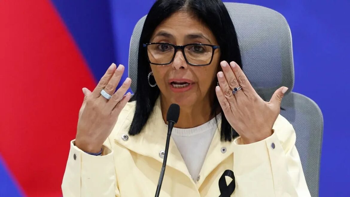 Woman wearing glasses and a light jacket speaks into a microphone, gesturing with both hands near her face during a press conference with a blue background.