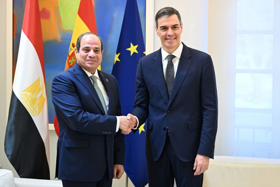 Two men in dark suits shake hands in a formal setting with flags (Egypt, Spain, EU) in the background.