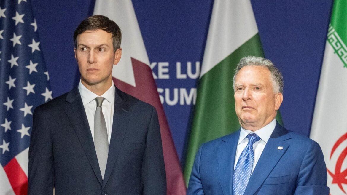 Two men in formal suits stand before a backdrop of flags at a diplomatic event, looking off to the side.