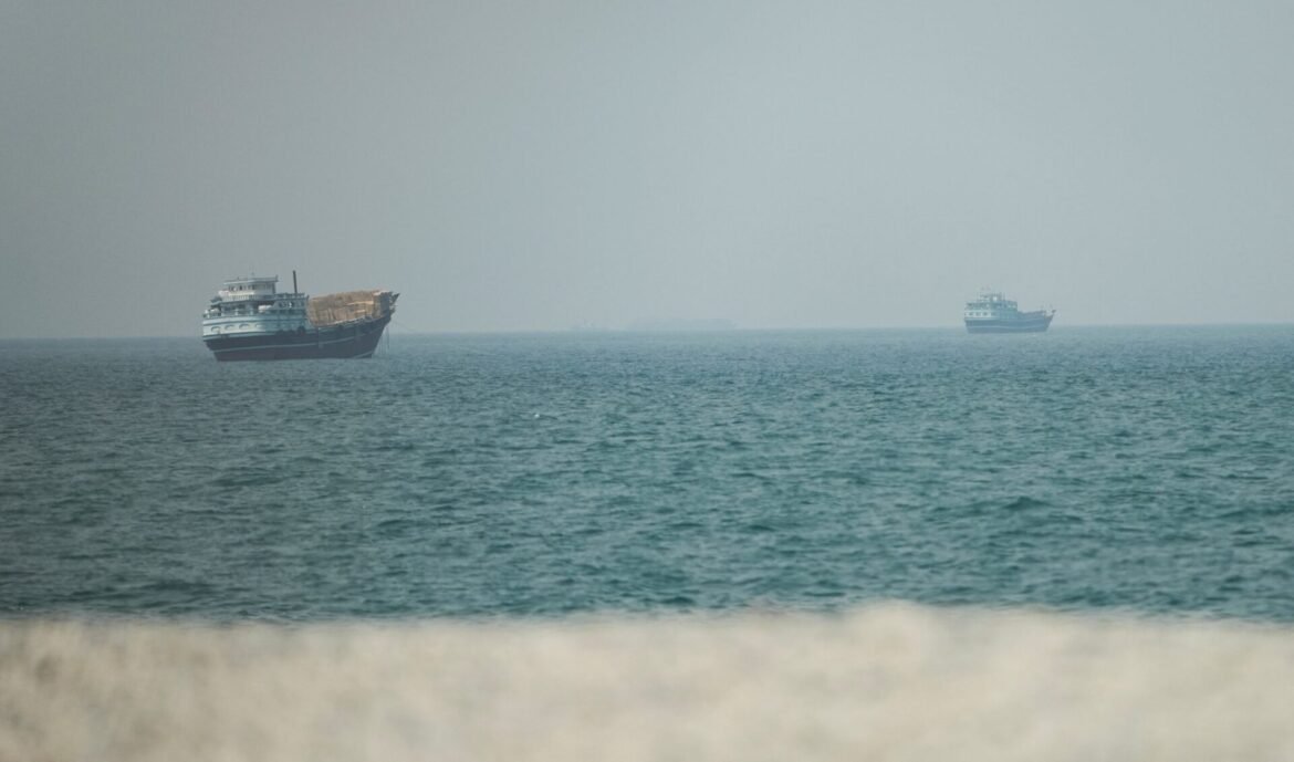 Two large ships on a calm blue sea, hazy horizon, with a sandy foreground.