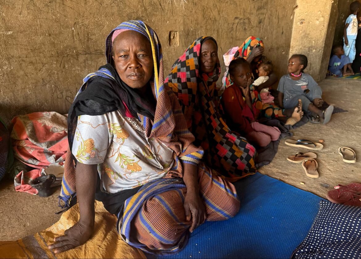 Group of women and children sitting on mats along a mud wall, wearing colorful traditional clothing and headscarves.