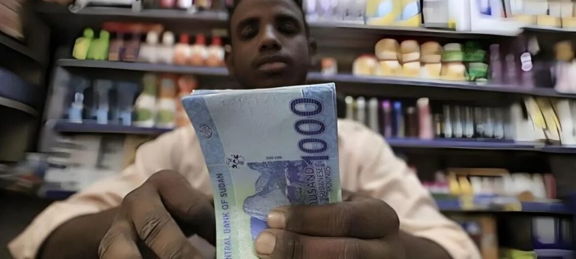 A man in a shop holding a stack of 1,000 Sudanese pounds toward the camera with shelves of products behind him.