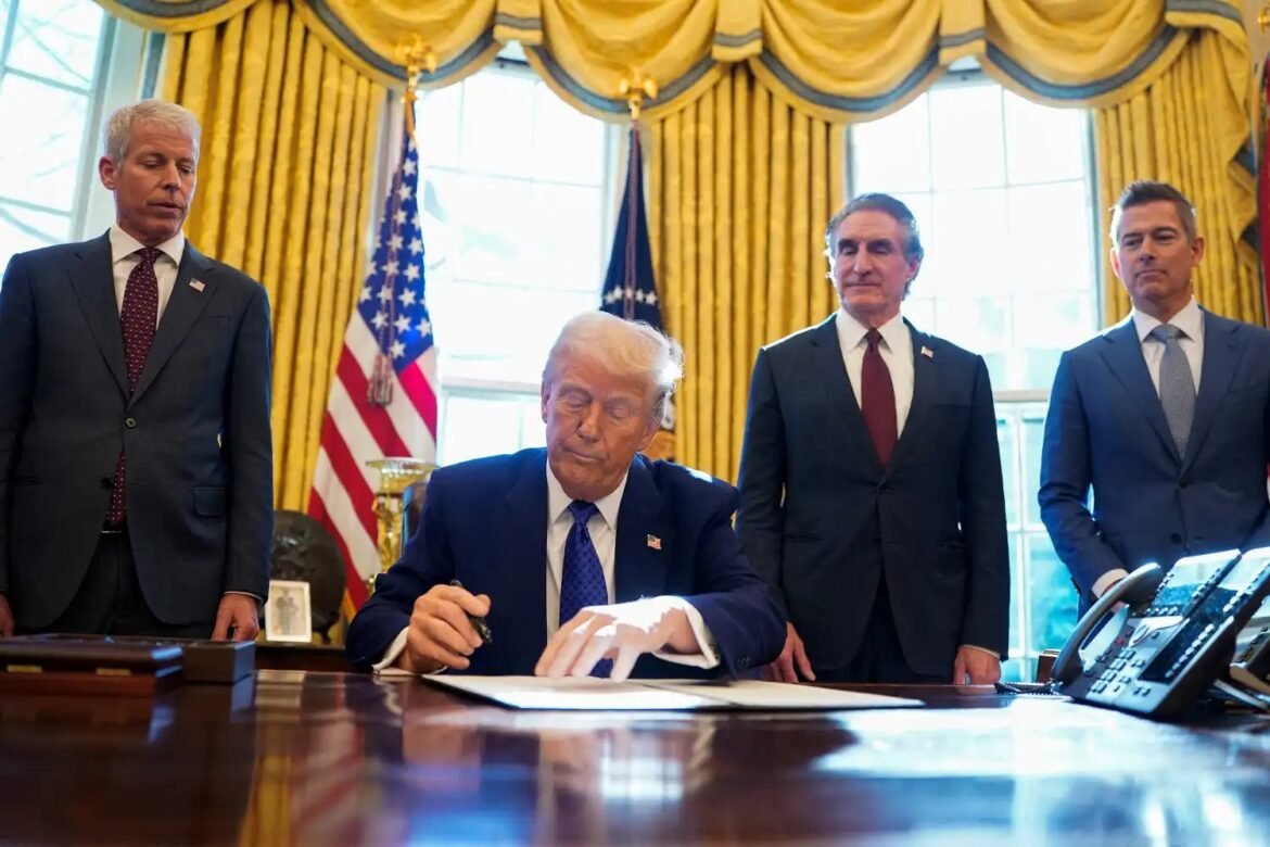 Former President Donald Trump signs a document at a desk in a gold-curtained room, flanked by three men in suits with American flags in the background.