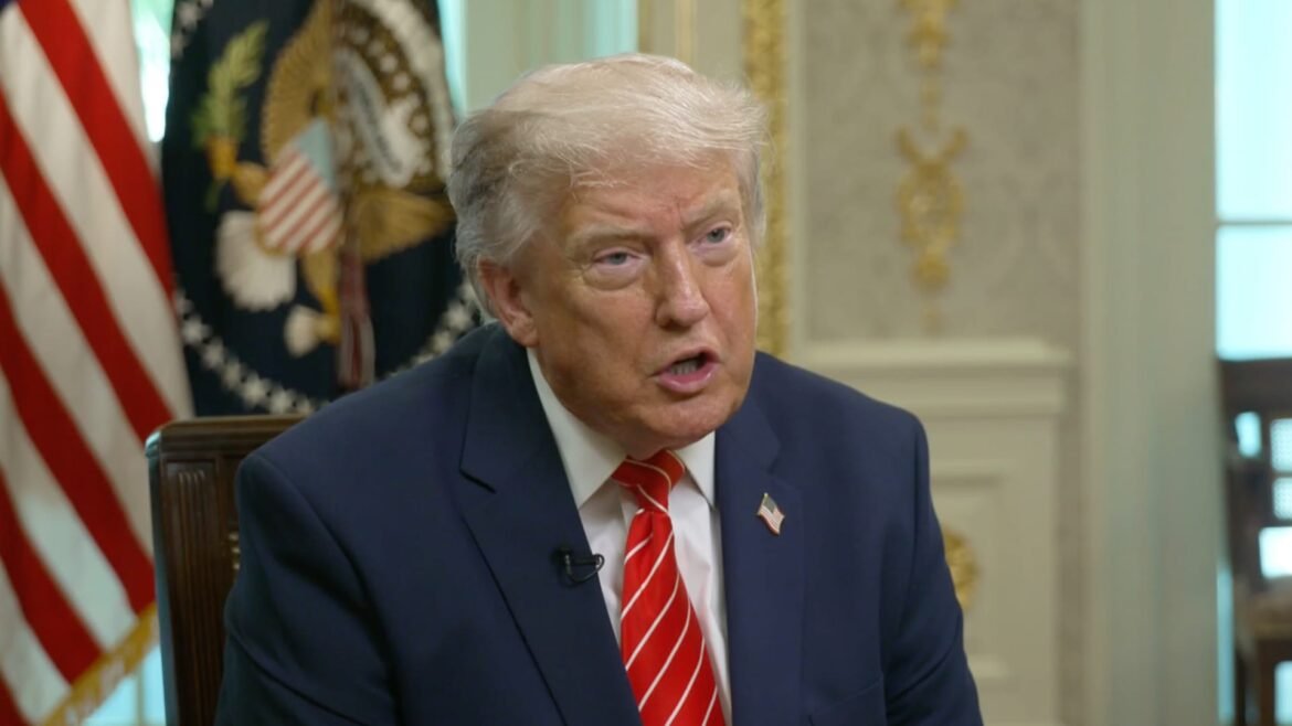 Man in a navy suit with a red striped tie speaks in a formal room with flags and ornate decor behind him (interview setting).
