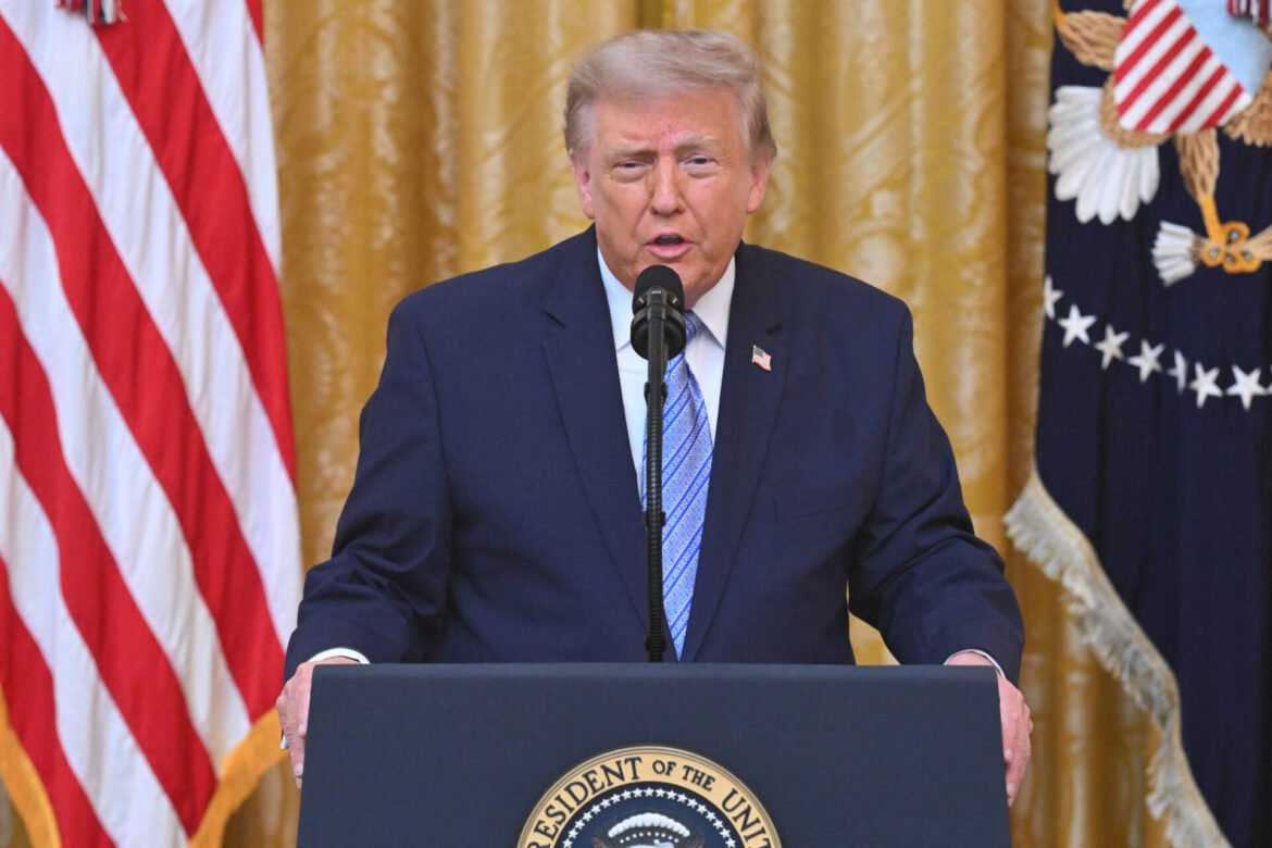 Man in a dark suit delivering a speech at a podium with the presidential seal, flanked by U.S. flags and a gold curtain backdrop.