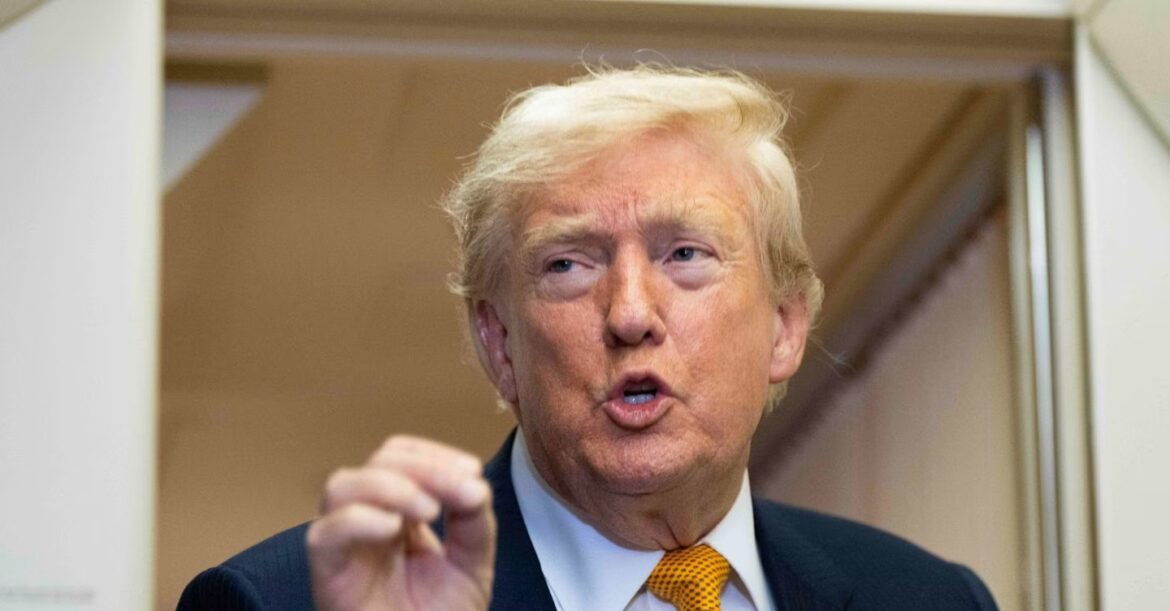 Man in a dark suit and orange tie speaking, mouth open, hand raised, in a doorway near camera edge.