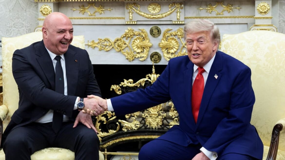 Two men in formal suits shake hands and smile while seated on gold-trimmed chairs in an ornate room.