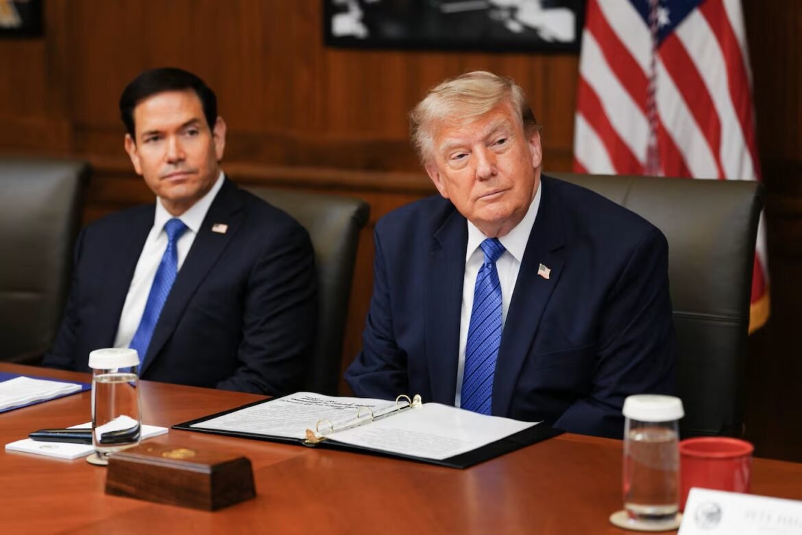 Two men in dark suits sit at a wooden conference table with an open binder and documents, an American flag behind them.