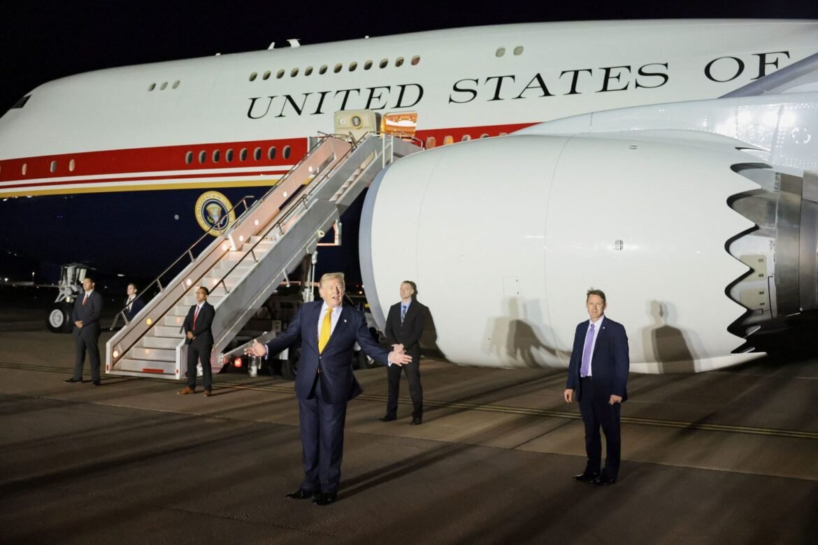 A man in a dark suit gestures with arms spread, standing on a tarmac beside a large white government airplane with 'UNITED STATES' on the fuselage and a mobile stairway nearby.