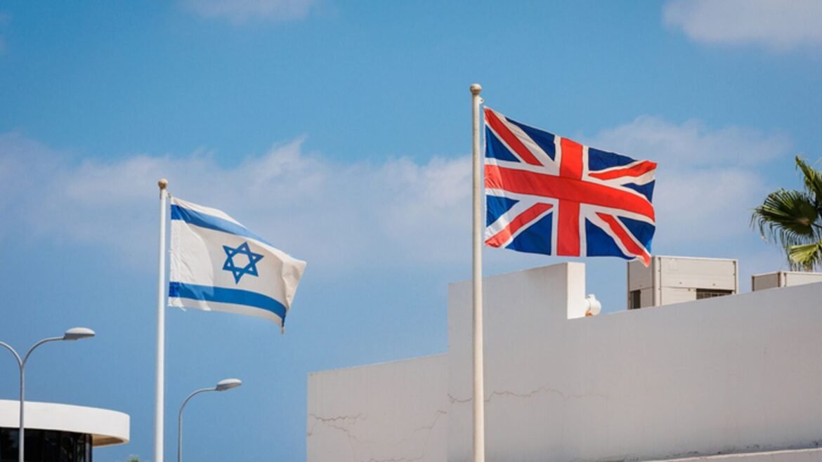 Union Jack and Israeli flag fluttering on flagpoles against a bright blue sky above a white building.
