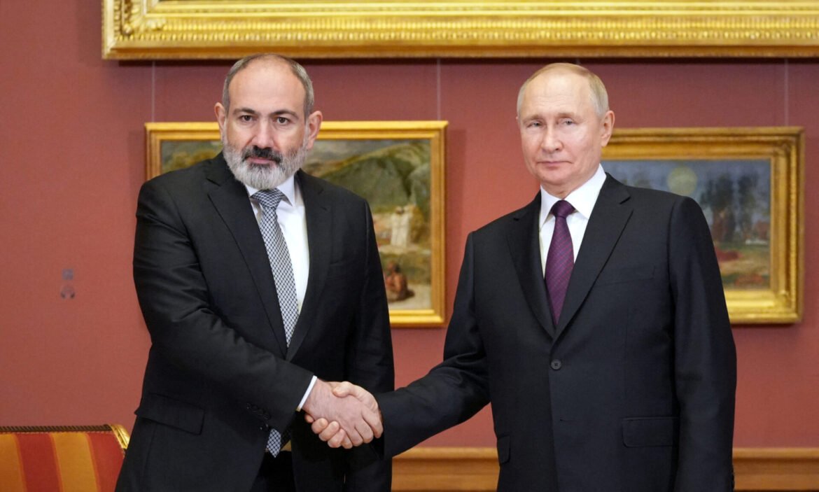 Two men in dark suits shake hands in a formal room with red walls and framed paintings behind them.