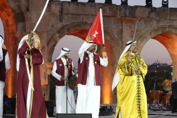 Group of men in traditional robes performing a sword dance on an outdoor stage with ancient stone arches at dusk; one raises a sword and flag.