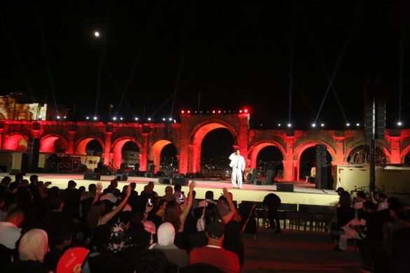 Nighttime outdoor concert at an arched stone venue with red lighting; a singer in white performs on stage as the audience watches.