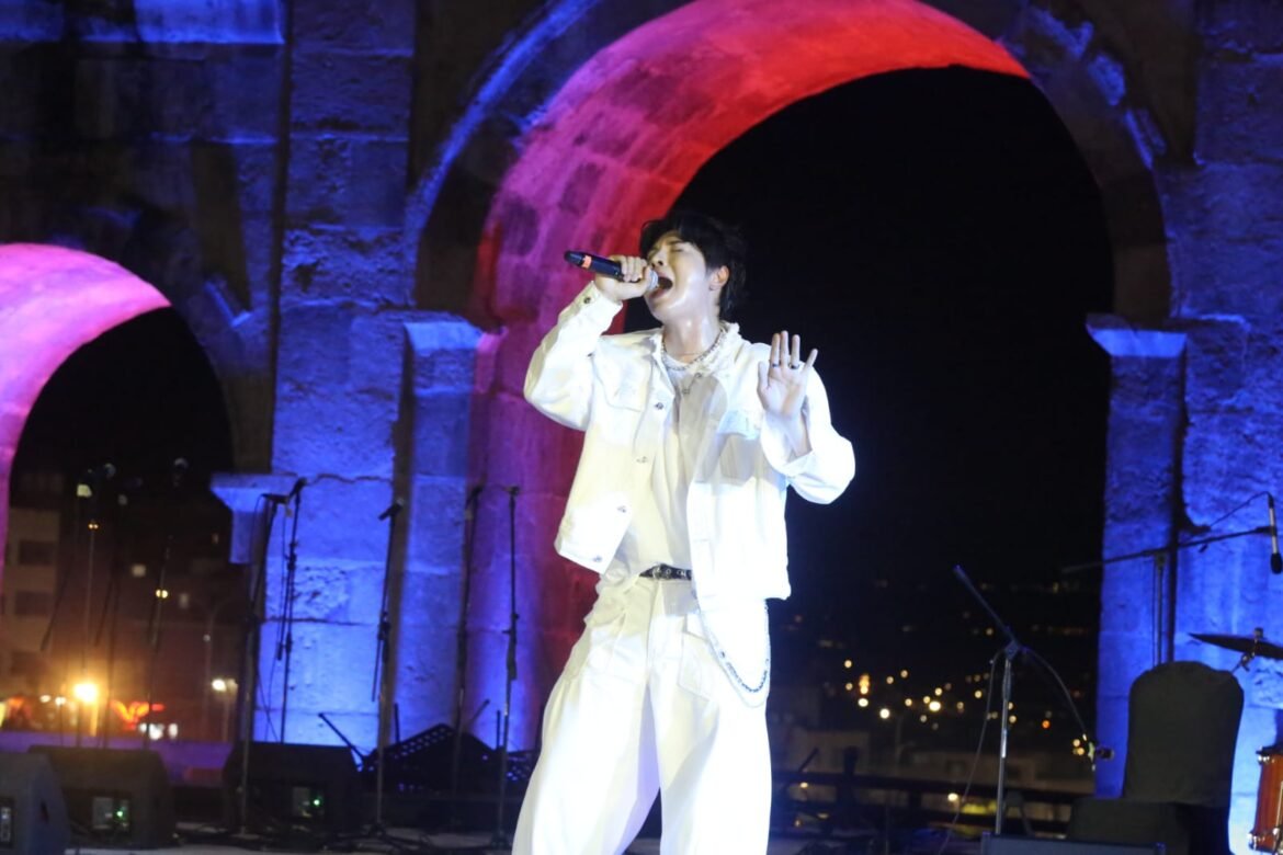 Male singer in white outfit performing on an outdoor stage at night, microphone in hand, colorful illuminated arches behind him.