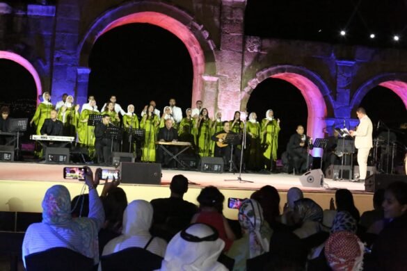 Choir in green dresses and white headscarves performs on an illuminated outdoor stage with arches; a conductor in a light suit leads the musicians.
