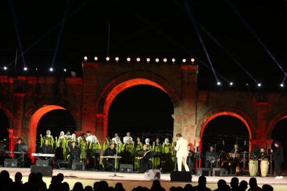Choir in green dresses performs on a historic stone arch stage at night, with a conductor in a light suit and a live band under red lighting.