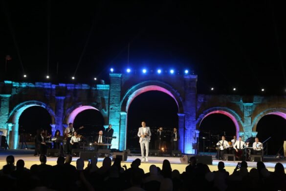 Nighttime outdoor concert on a stone arch stage; a singer in a light suit performs while a full band plays under blue lights.