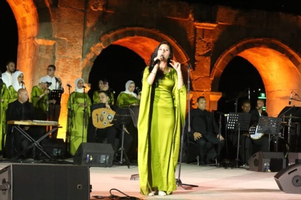 Female singer in a bright lime-green gown performs on an outdoor stage with a gold-toned stone arch backdrop and a live orchestra behind her.