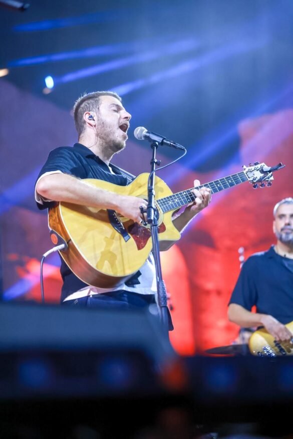 Male musician singing into a microphone while playing a yellow acoustic guitar on stage under colorful lights in concert.