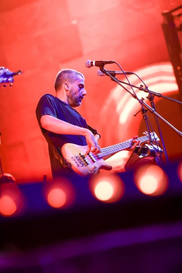 Male musician playing an electric bass on stage under red lights, singing into a microphone stand nearby