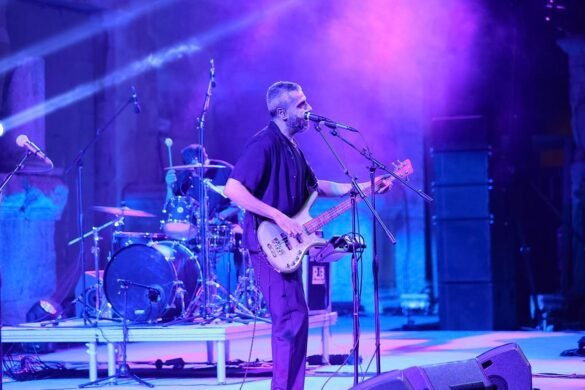 Male bassist sings into a microphone while playing a bass on a blue-lit stage with a drummer in the background