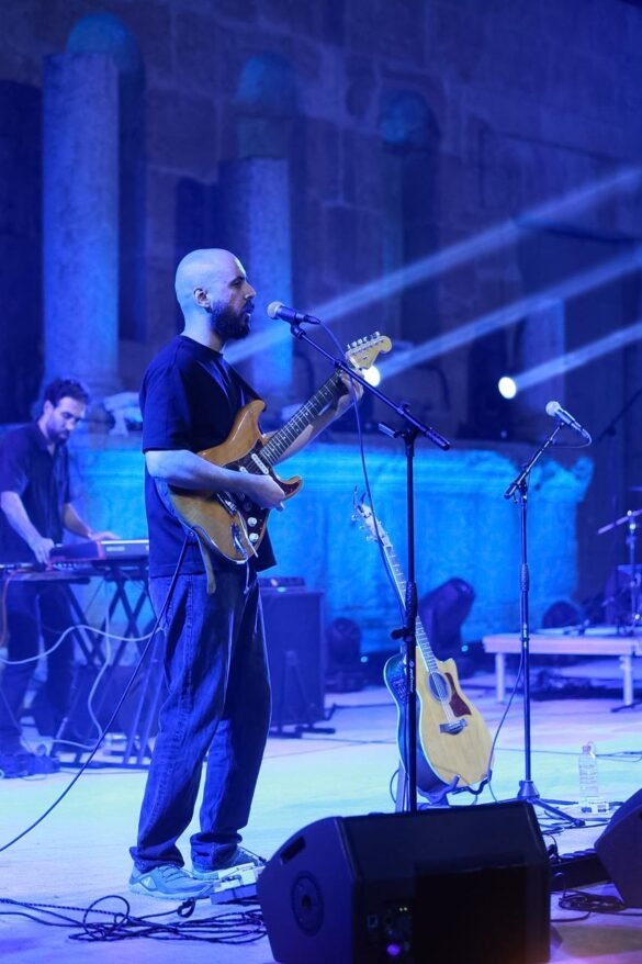 A bearded man playing an electric guitar and singing into a microphone on a blue-lit stage, with a keyboardist in the background.