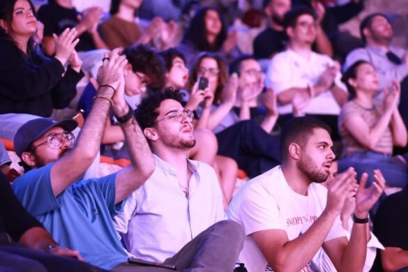 Audience at a live show applauds, with a man in a blue cap and glasses and another man beside him in the foreground clapping.