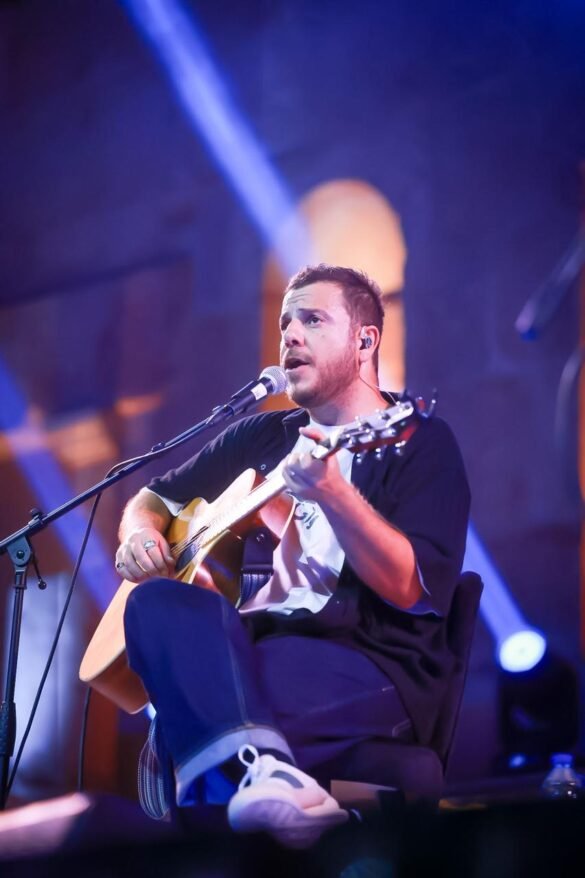 Male musician sitting on a chair, playing an acoustic guitar and singing into a microphone on stage.