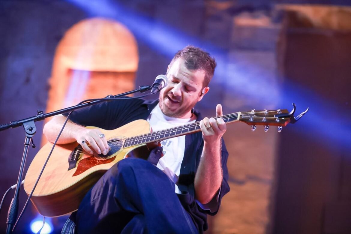 Male guitarist performing live on stage, singing into a microphone while playing an acoustic guitar under blue stage lights