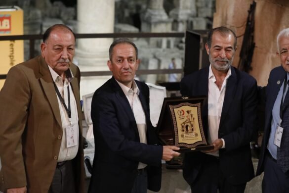 Four men in suits pose with an ornate award plaque featuring an arch, as others look on in a ceremony setting.