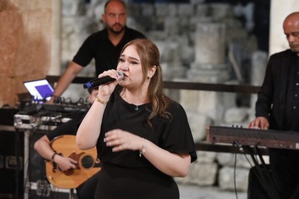 Female singer in a black outfit sings into a mic, with musicians and stage equipment in the background.