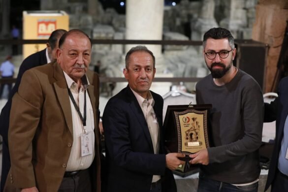 Three men pose with an award plaque during a formal event, ancient ruins visible in the background.