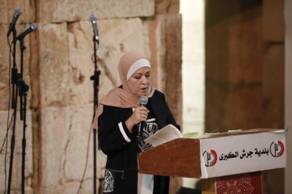 Woman in a peach hijab speaks into a handheld microphone at a wooden podium with a banner, in a stone-walled setting.