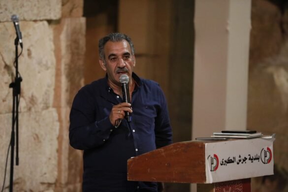Man in a dark blue shirt speaks into a handheld microphone at a wooden podium, with Arabic banner visible on the front.