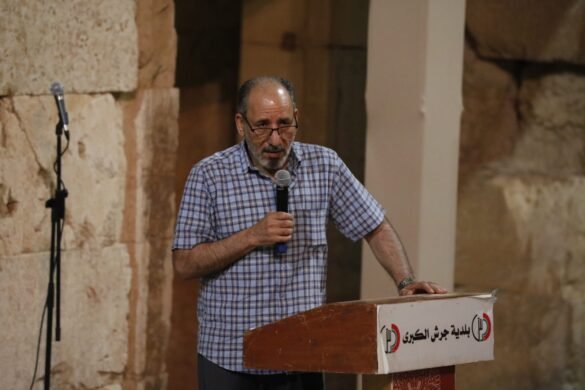 Middle-aged man with glasses speaks into a handheld microphone at a wooden podium in a stone-walled venue