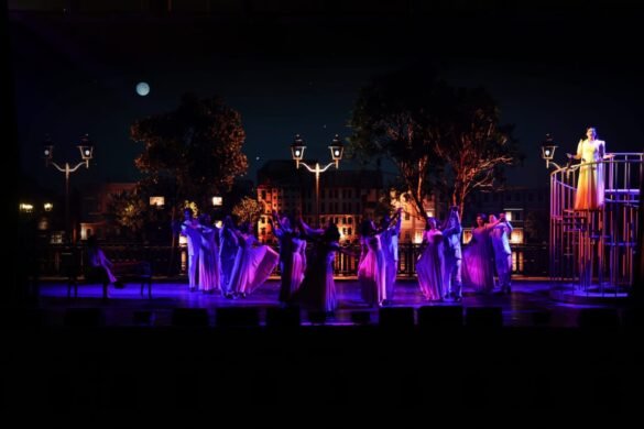 Night outdoor stage performance with dancers in lavender gowns and a performer in a yellow dress on an elevated platform against a city backdrop and moon.