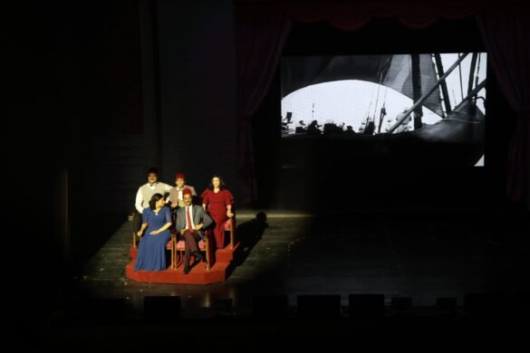 Six actors sit on a red platform on a dark theatre stage, dressed in formal clothes, performing with a large black-and-white ship scene projected behind them.