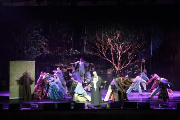 A large group of performers in colorful robes dances on a dimly lit stage with a bare tree and rain effects in the background.