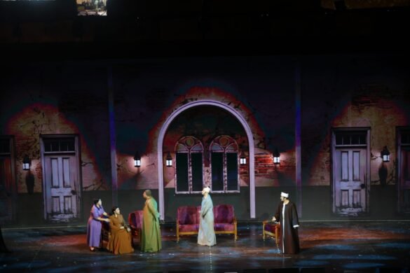 Theater stage scene: performers in period robes around a sofa on a cracked-wall set with arches and doors in the background.