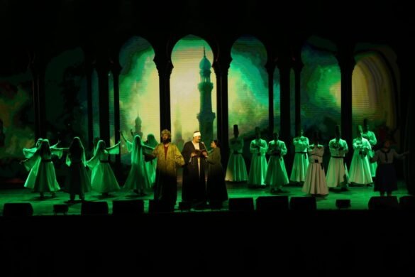 Group of performers on a stage under green lighting, wearing flowing white robes; central figures appear to be speaking or singing with a backdrop of archways and domed towers.