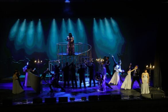 Performer in a gown stands on a spiral balcony center stage, surrounded by dancers in formal dresses under blue spotlights.