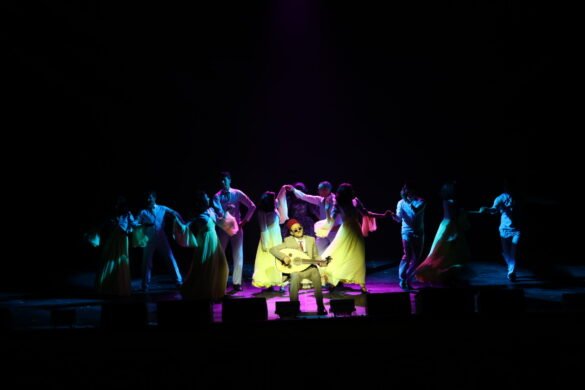 A musician in a suit sits center stage playing a guitar while colorful dancers sway around him under dramatic lighting on a dark concert stage.